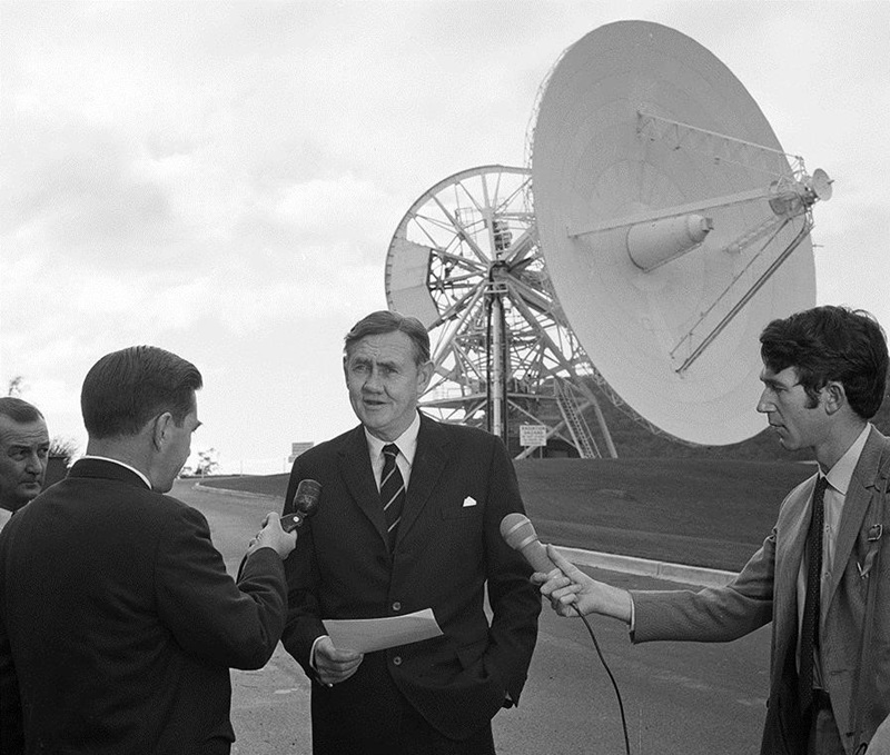 Prime Minister John Gorton standing in front of the Honeysuckle Creek Tracking Station telescope giving an interview to two reporters holding microphones. 