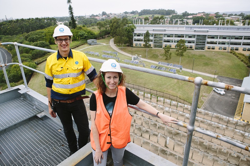 Two people (including Dr Jenny Hayward) standing on top of a building in high vis workwear, CSIRO branded merch with solar panels on the ground below them.