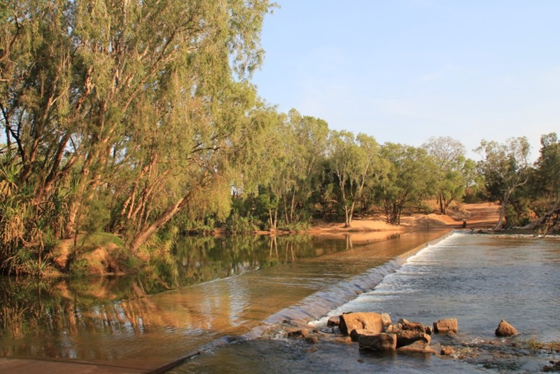 A river crossing with water flowing over a road and red sand and dirt on the other side.
