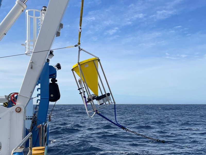 A piece of scientific equipment that looks like a large yellow cone hangs from a wire above the ocean.