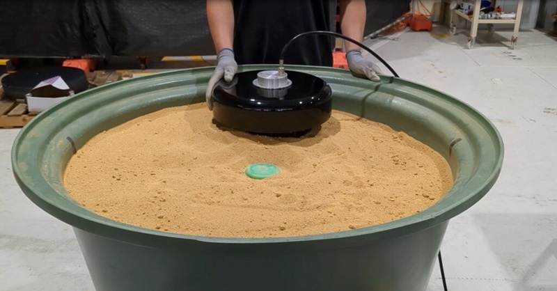 A photo showing a large tub of sand. Atop the sand is a round black metal object which is used to detect landmines and a person is holding the object.