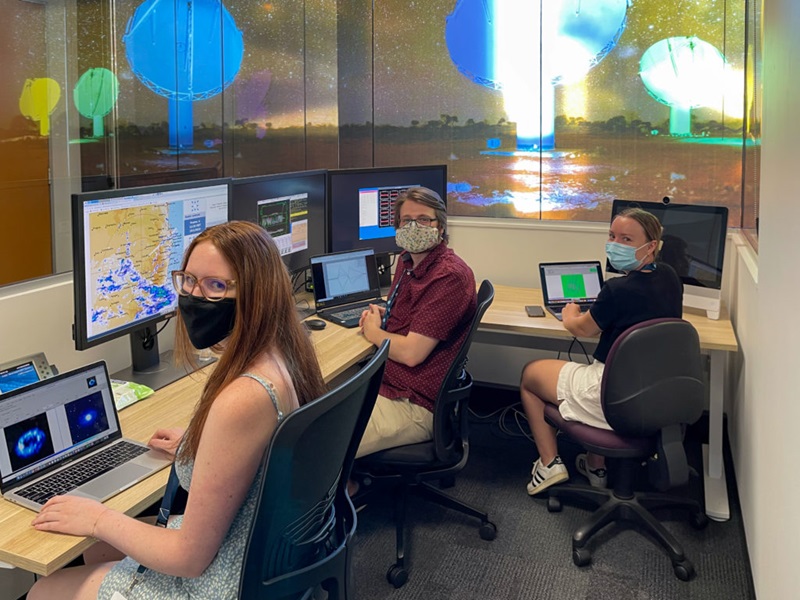 Three vacation student participants seated at computers in an office space wearing face masks