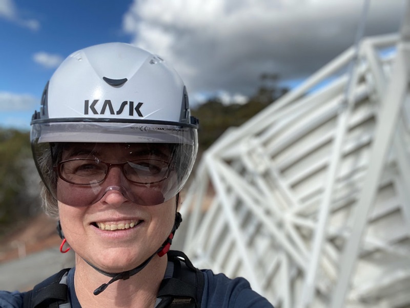 Photo of Suzy Jackson wearing a helmet, looking at the camera with a blue sky and machinery in the background.