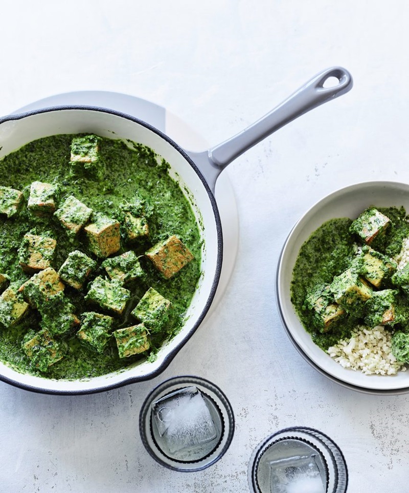 A pot sits on a stone top bench, inside it is tofu saag. Next to the pot are two glasses of iced water and a separate dish of tofu saag  