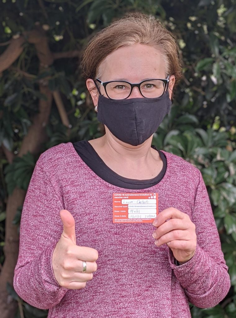 Woman wearing mask holds up her vaccine card and takes a vaccine selfie.