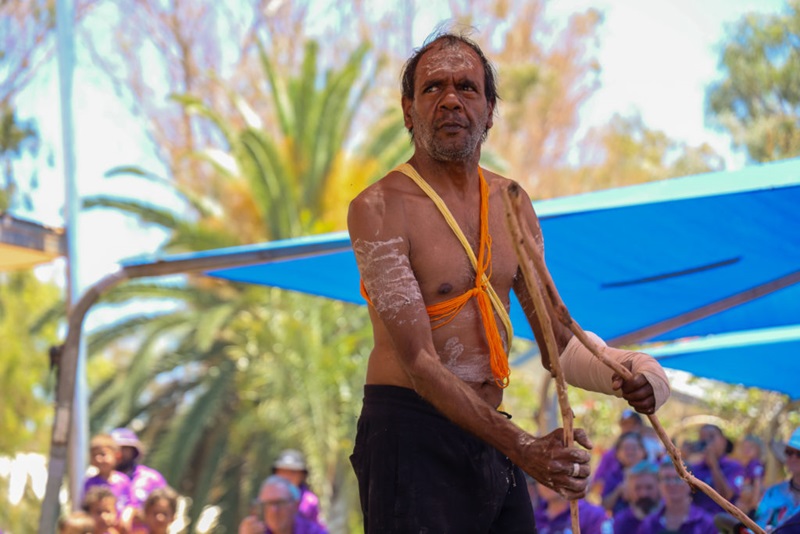 Wajarri man with ochre painted on his torso, arms and face. Yellow and orange threads are tied around his body. His left arm is bandaged and he is holding two long sticks. He is performing a traditional dance.