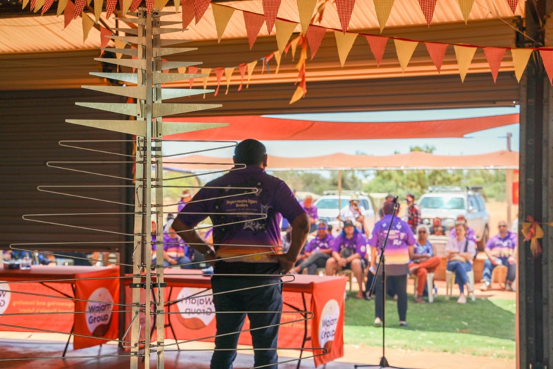 A metal frame in the shape of a tree can be seen in the foreground. Behind the metal frame, Des Mongoo can be seen with his back to the camera. He is wearing a purple "Inyarrimanha Ilgari Bundara" shirt. In front of him are 2 tables with orange covers which say "Wajarri Group", and next to the tables is a microphone stand. Behind the tables a group of people, most of whom are wearing the same purple shirts, can be seen seated under a shade.