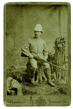 Old image from the 1800s of a man sitting on a tree stump holding a rifle across his thighs. 