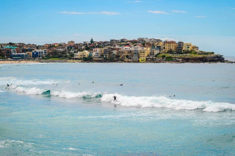 A beach with surfers riding a wave into shore and buildings on a hill behind the cove.