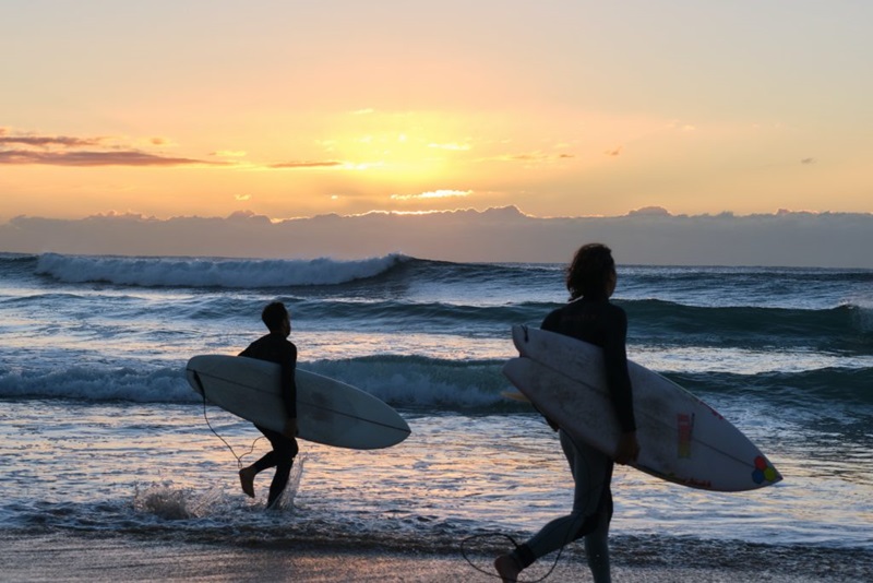 Two male surfers head into the ocean with boards under their arms at sunset. 