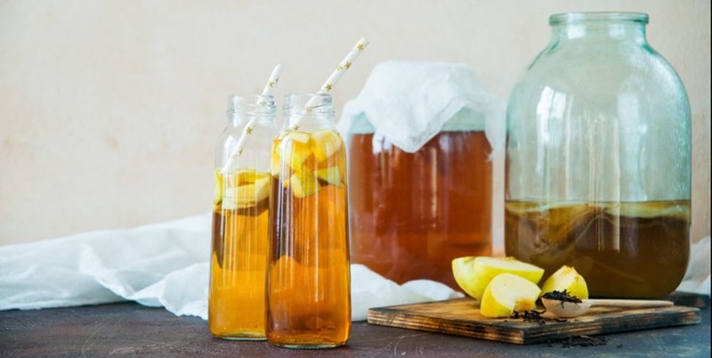 Two vials of orange looking liquid with straws in the foreground. There are jars with the same orange liquid in the background with cloth over it.