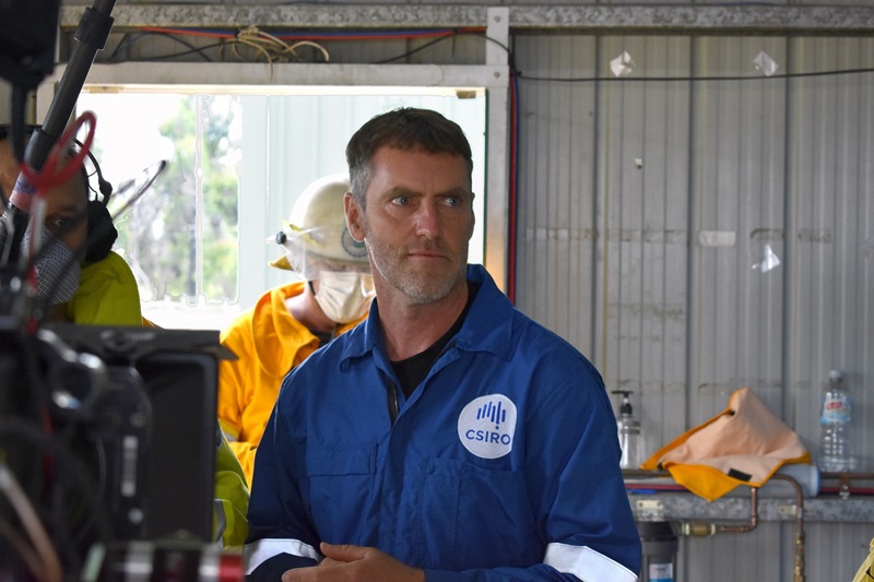 A person wearing bright blue coveralls standing in a shed looking to the right.