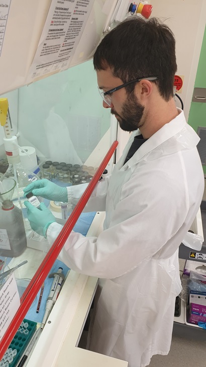 A person in a white lab boat and blue-green gloves working with bottles and jars in a fume hood.