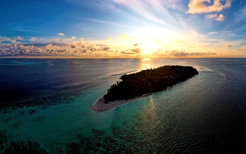 Aerial image of a dark island surrounded by coral reefs in the blue ocean. The sun is low in the sky behind the island.