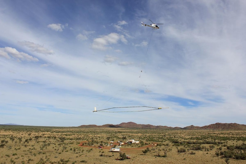 A helicopter with a large ring suspended from it hovering over landscape