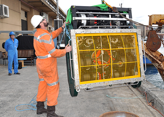 Man in orange overalls helping load a big instrument with a crane