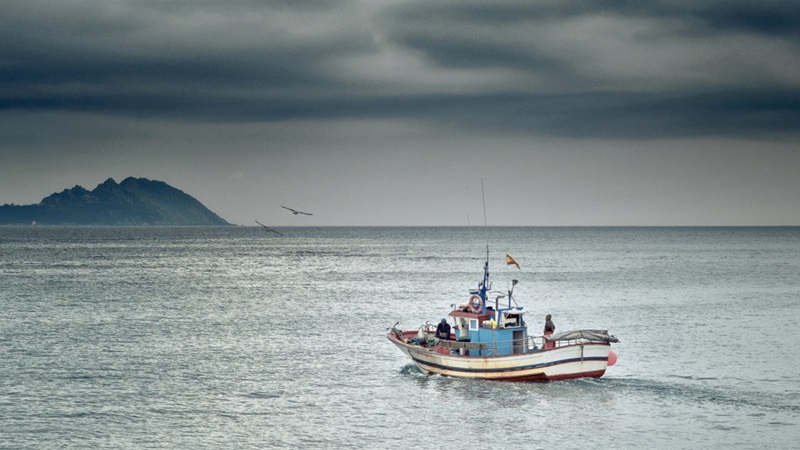 Fishing boat on grey ocean waters with sea gulls circling against a grey, gloomy sky. 
