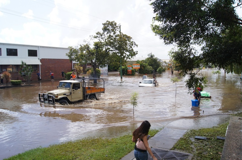 A flooded street with people and cars trying to navigate through waist-high brown water. 