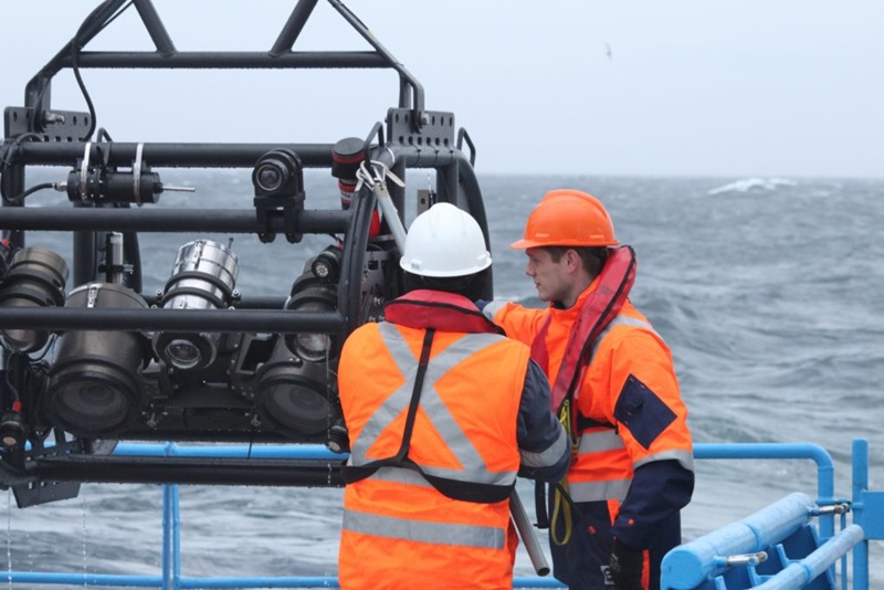 two men in hard hats and high vis pulling a piece of equipment on board with choppy sea in the backhround