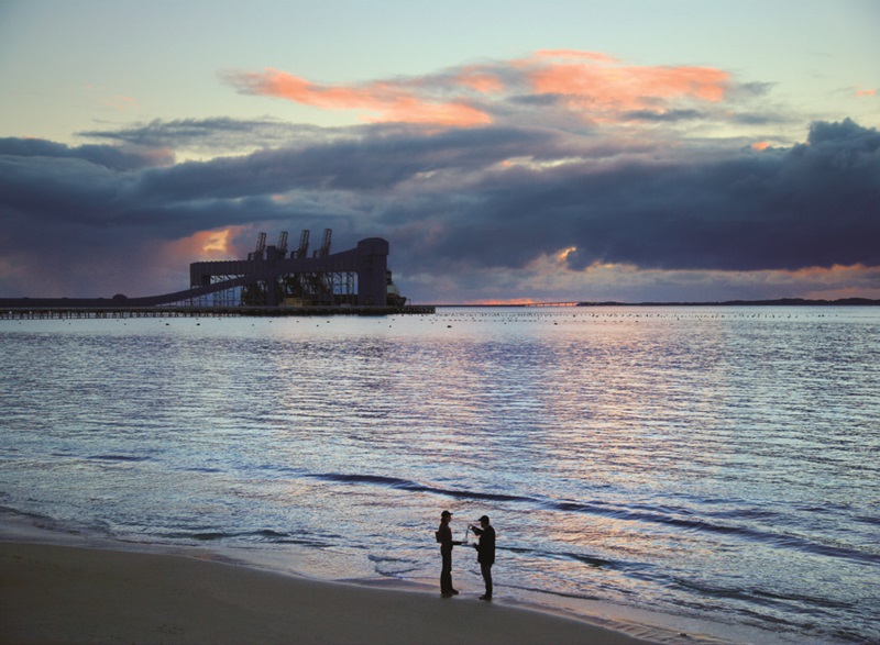 Two people standing on a beach with water and industry in background