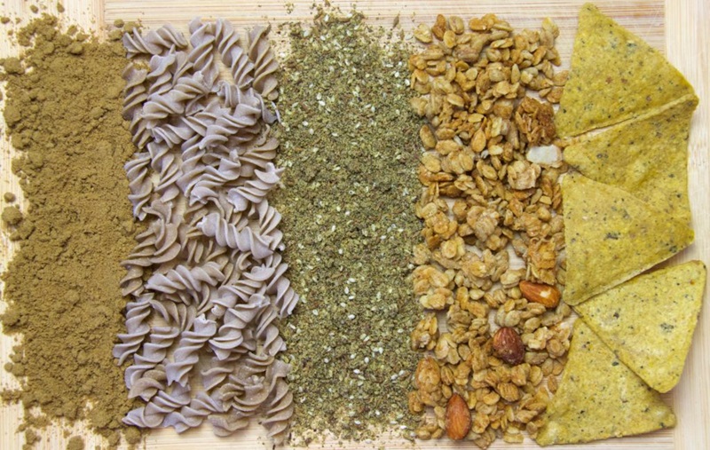 A picture of 5 different substances on a woodden table: cricket flour, pasta, dukkha, granola and chips. All of them have cricket flour in them. 