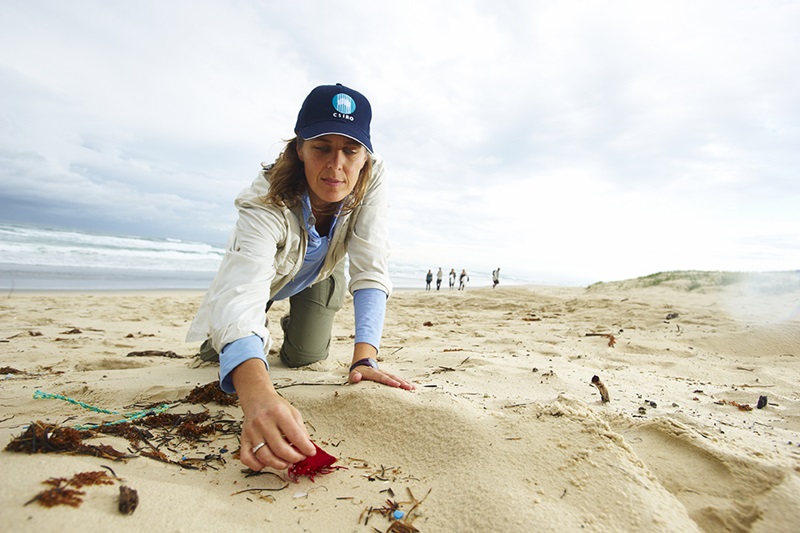 A woman on a beach picking up a piece of red plastic from sand