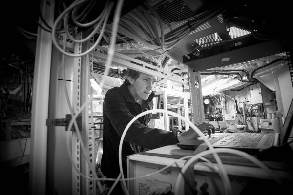 black and white photo close up of man on laptop surrounded by shelves and equipment and wires