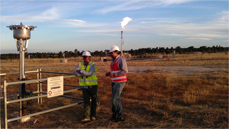 Two mean leaning on fence next to a bore coming out of ground and gas well in background with flames emerging