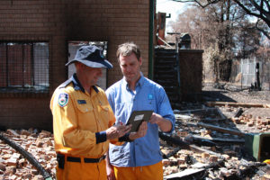 Two men looking at a tablet device with burnt houses in background