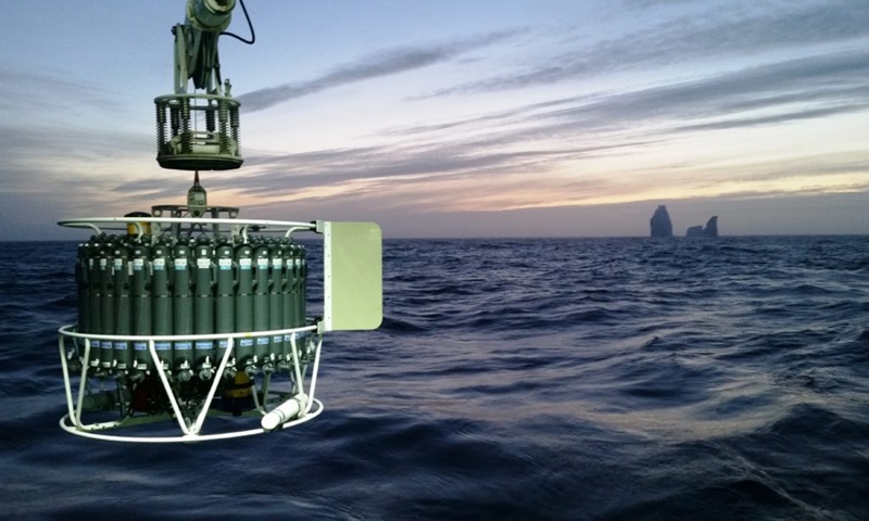 A large circular scientific instrument hangs over the ocean with an iceberg in the background.