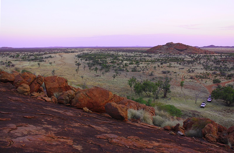 A landscape with red rocky mountains and grassy plains with two vehicles in view at base of mountain