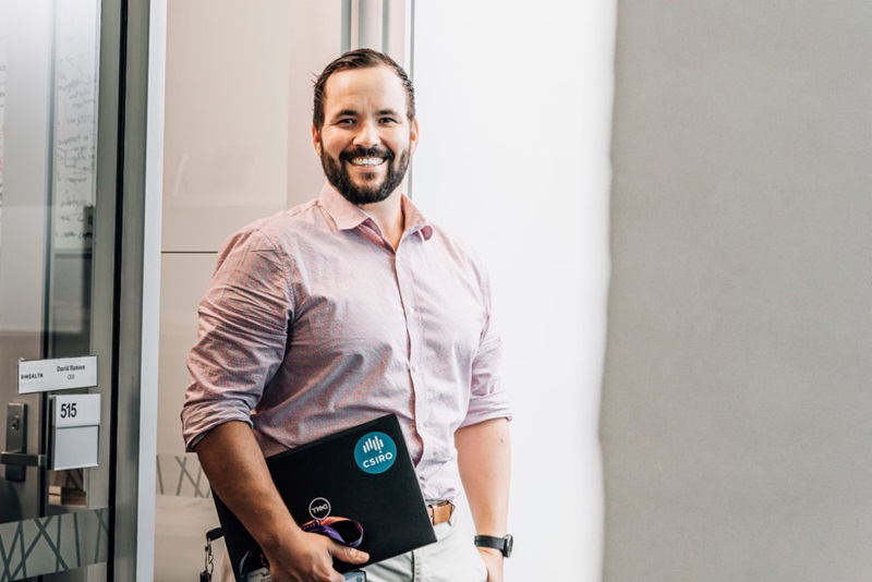 A man stands in a door way, smiling, hodling a laptop.