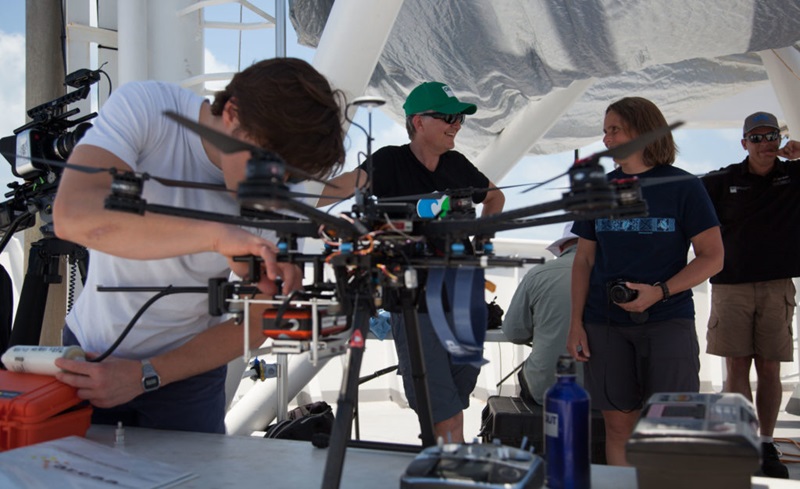 A scientist works on an aerial drone while three people stand on the deck of a ship behind them.