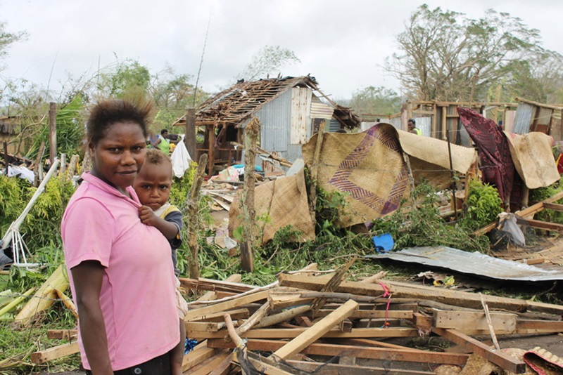 A woman holding a baby in front of a cyclone-damaged house in Vanuatu