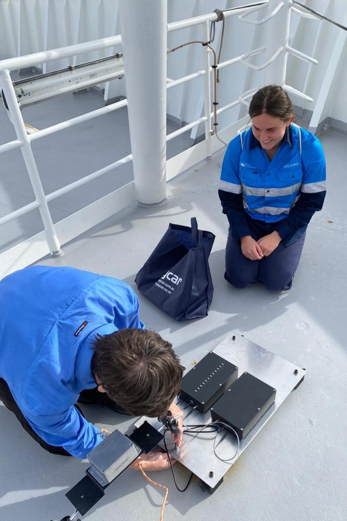 Two people on the deck of a ship kneeling over a piece of electronic equipment.