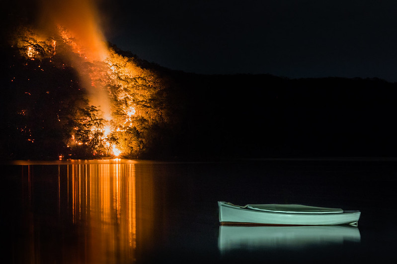 A boat sits on the water while the bush behind it (on the edge of the lake) is on fire.