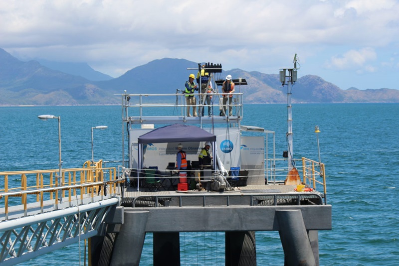 men working on equipment installed at the end of a jetty with ocean and island in the background