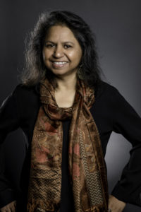 Dr Anu Kumar smiling at the camera against a grey background.