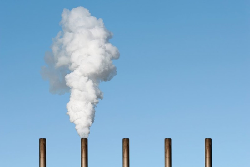 Top of five smoke stacks against a blue sky with one belching white clouds of smoke. 