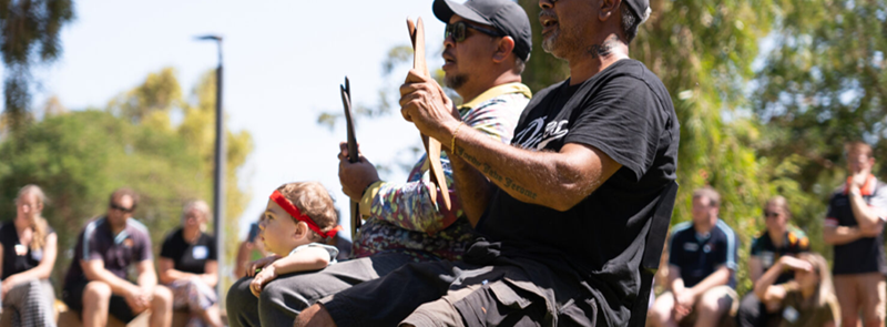Two Aboriginal men and a small boy watch dancers give a traditional Ngarluma Country welcome session, clapping time with sticks at Living STEM Showcase 2023 