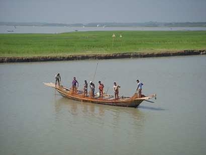 7 men stand in a long narrow boat with a fishing net. Rice paddies in the background