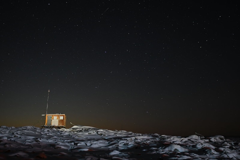 A small wooden hut perched on top of an icy hill, with the night sky behind.
