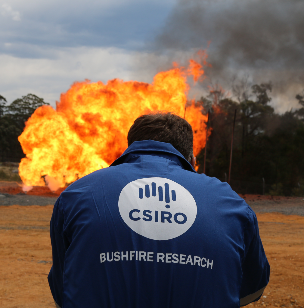 CSIRO scientist wearing a branded jacket, standing in front of flames and bushland.