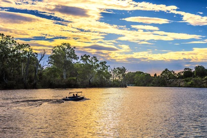 Sunset behind a river, with a small boat zipping along the foreground. Leafy gums on the river embankment.