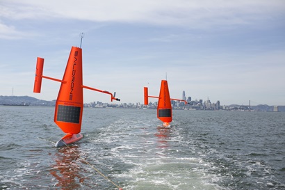 Two Saildrones are sailing in the water. A city can be seen in the distance.